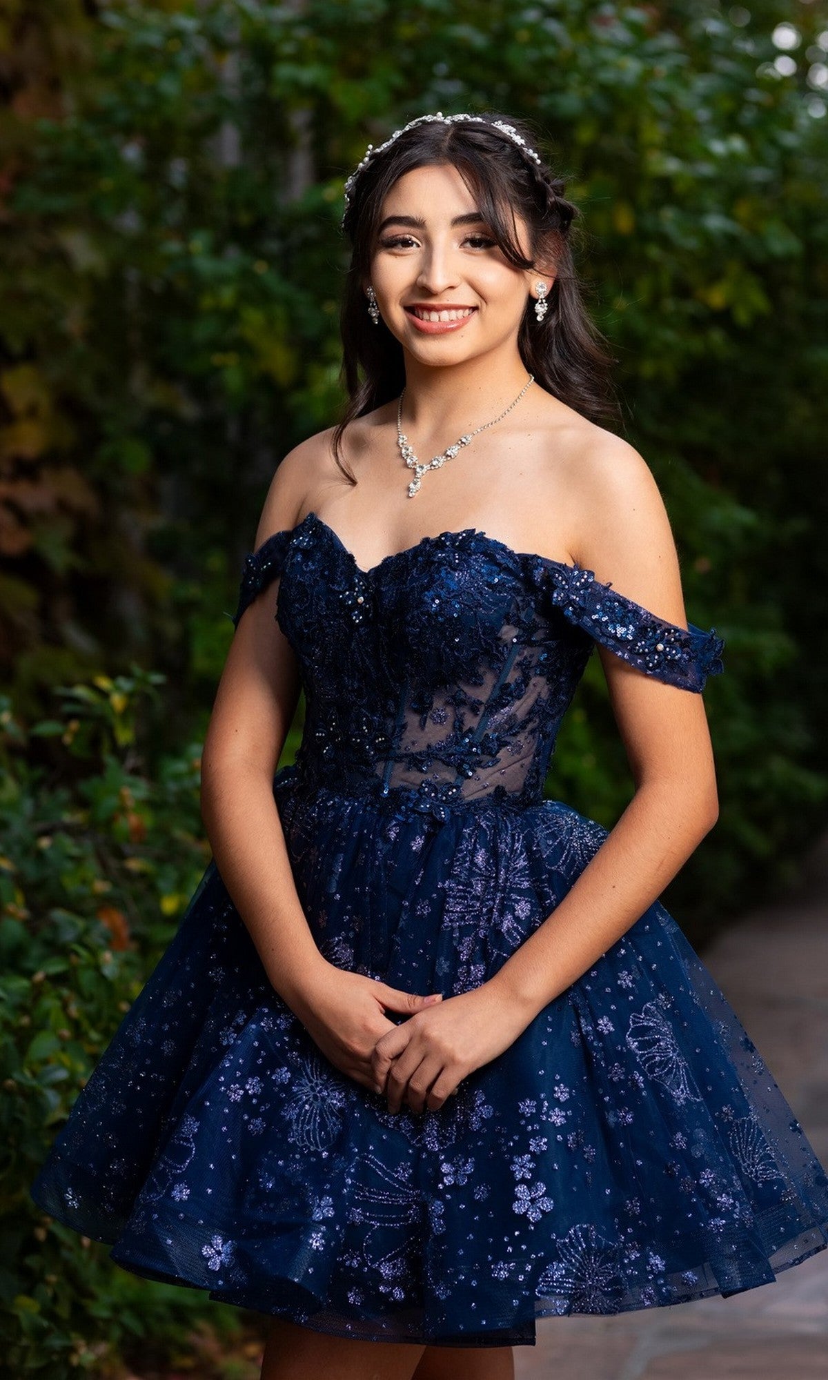 A young woman stands outdoors wearing the Chicas C377 Short Homecoming Dress in navy blue, adorned with floral patterns. The dress features a delicate tulle skirt and an elegantly embroidered off-the-shoulder corset bodice. She accessorizes with a necklace and a headpiece, smiling warmly at the camera as green foliage provides a lush backdrop.
